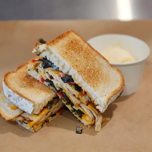 Toasted sandwich with melted cheese, black olives, and red peppers, served with a side of sauce.