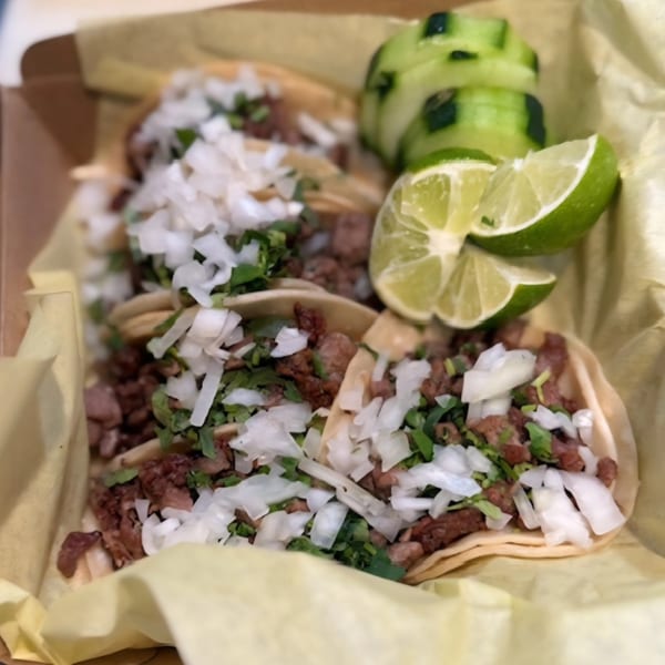Three Dank Donuts tacos with meat, onions, cilantro, lime wedges, and cucumber slices in a box.