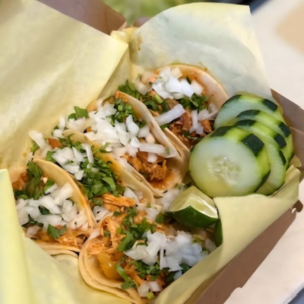 A box of tacos with shredded chicken, diced onions, cilantro, lime wedges, and sliced cucumbers.