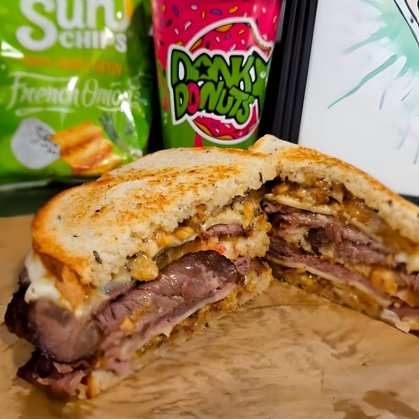 A sliced roast beef sandwich with melted cheese and caramelized onions, next to a Dank Donuts cup.