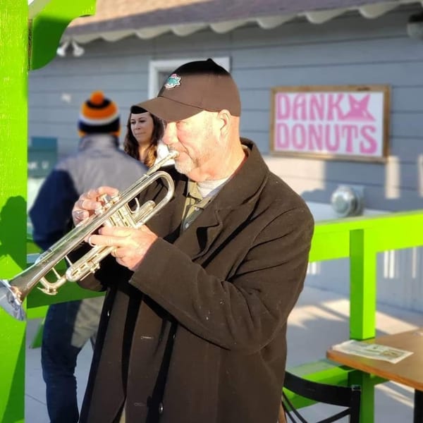 A man in a black coat and cap plays a trumpet outside Dank Donuts.