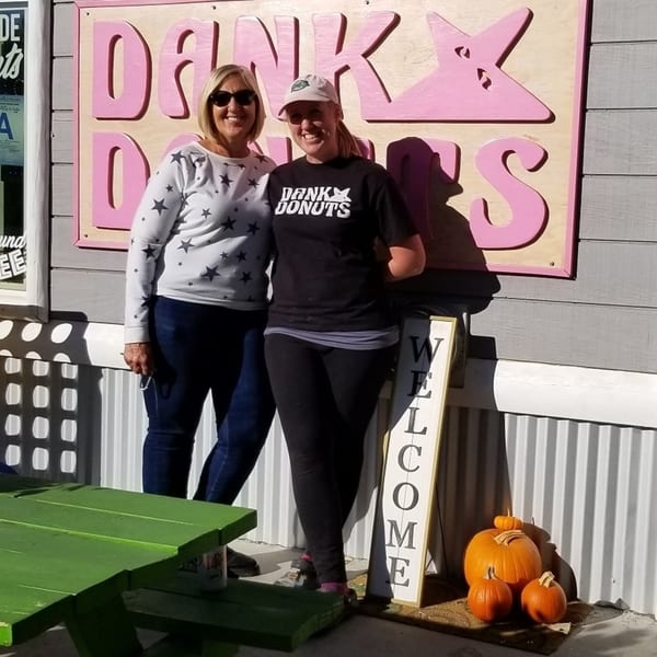 Two women smiling in front of the Dank Donuts sign, with pumpkins and a green picnic table.