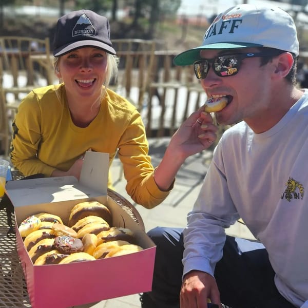 A woman feeds a man a Dank Donut from a pink box on an outdoor patio.