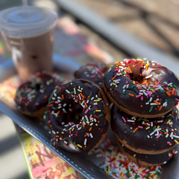 Chocolate-frosted Dank Donuts with colorful sprinkles on a tray, with an iced coffee in the background.