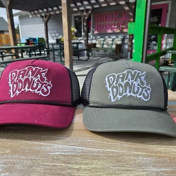 Two Dank Donuts trucker hats, one magenta and one gray, on a wooden table outdoors.