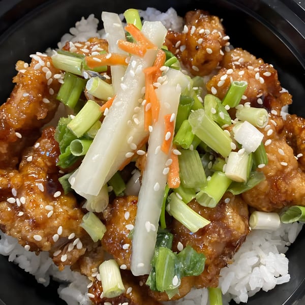 Close-up of a rice bowl with glazed chicken, green onions, pickled daikon, and carrots.