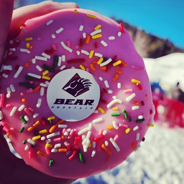 Pink frosted donut with colorful sprinkles and a Bear Mountain logo, held against a snowy mountain background.