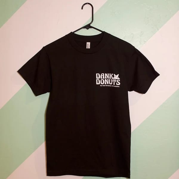 Black Dank Donuts t-shirt with white logo hanging on a black hanger against a mint green and white wall.