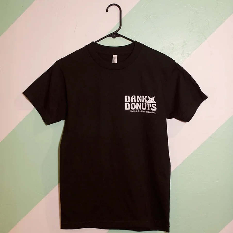 Black Dank Donuts t-shirt with white logo hanging on a black hanger against a mint green and white wall.