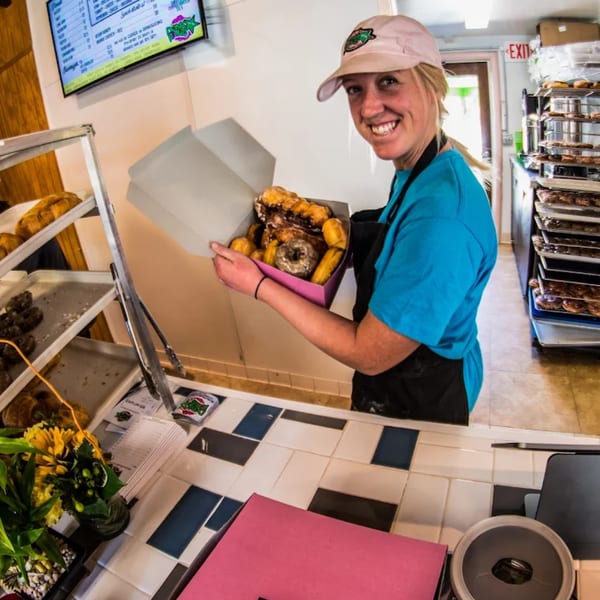 A smiling Dank Donuts employee in a cap and apron holds a box of various donuts.