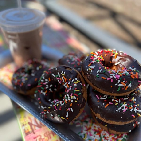 A tray of Dank Donuts' chocolate-frosted donuts with colorful sprinkles, alongside an iced coffee.