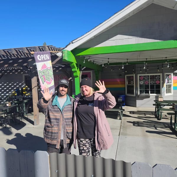 Two smiling Dank Donuts employees wave outside the colorful donut shop on a sunny day.